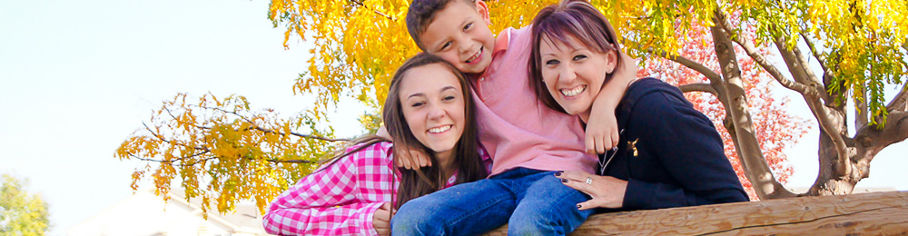 Mother with teenage daughter and young son smiling at the camera with a yellow tree behind them