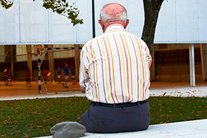 back of an elderly man sitting on a bench looking towards a building