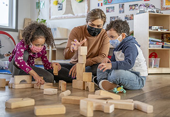 A teacher helps daycare kids build a structure with wooden blocks on the floor.