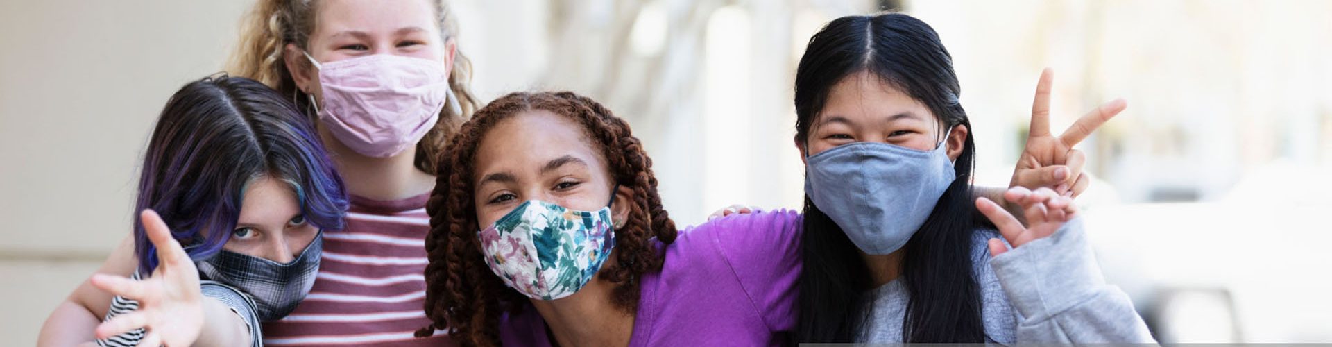 A multi-ethnic group of four tween girls, 12 and 13 years old, standing together on a sidewalk outside a building. They are wearing protective face masks during the COVID-19 pandemic, trying to prevent the spread of coronavirus. They are looking at the camera, smiling behind their masks.