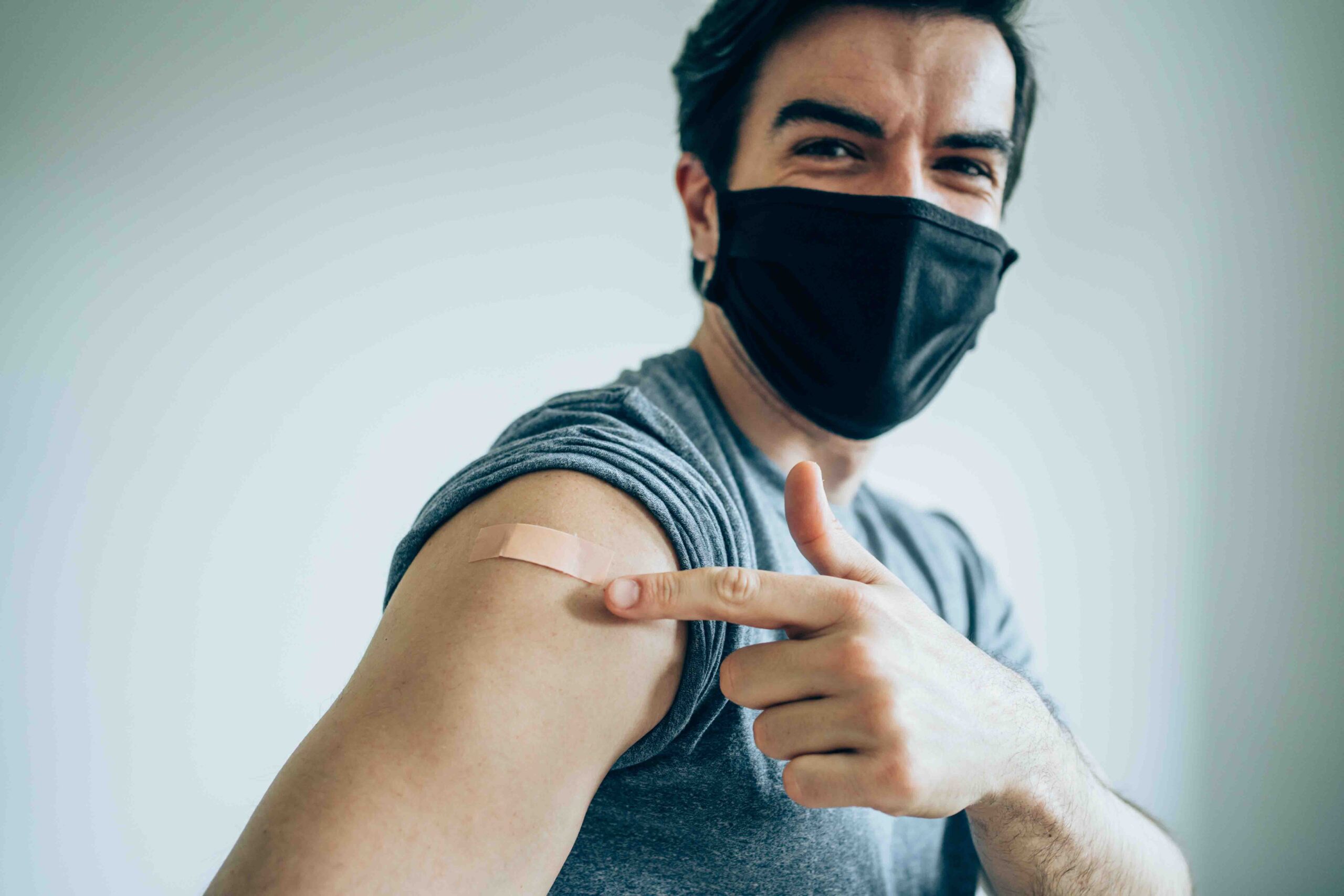 Man wearing protective face mask and pointing at his arm with a bandage after receiving COVID-19 vaccine. Young man showing his shoulder after getting coronavirus vaccine at vaccination center. Focus is on the bandage.