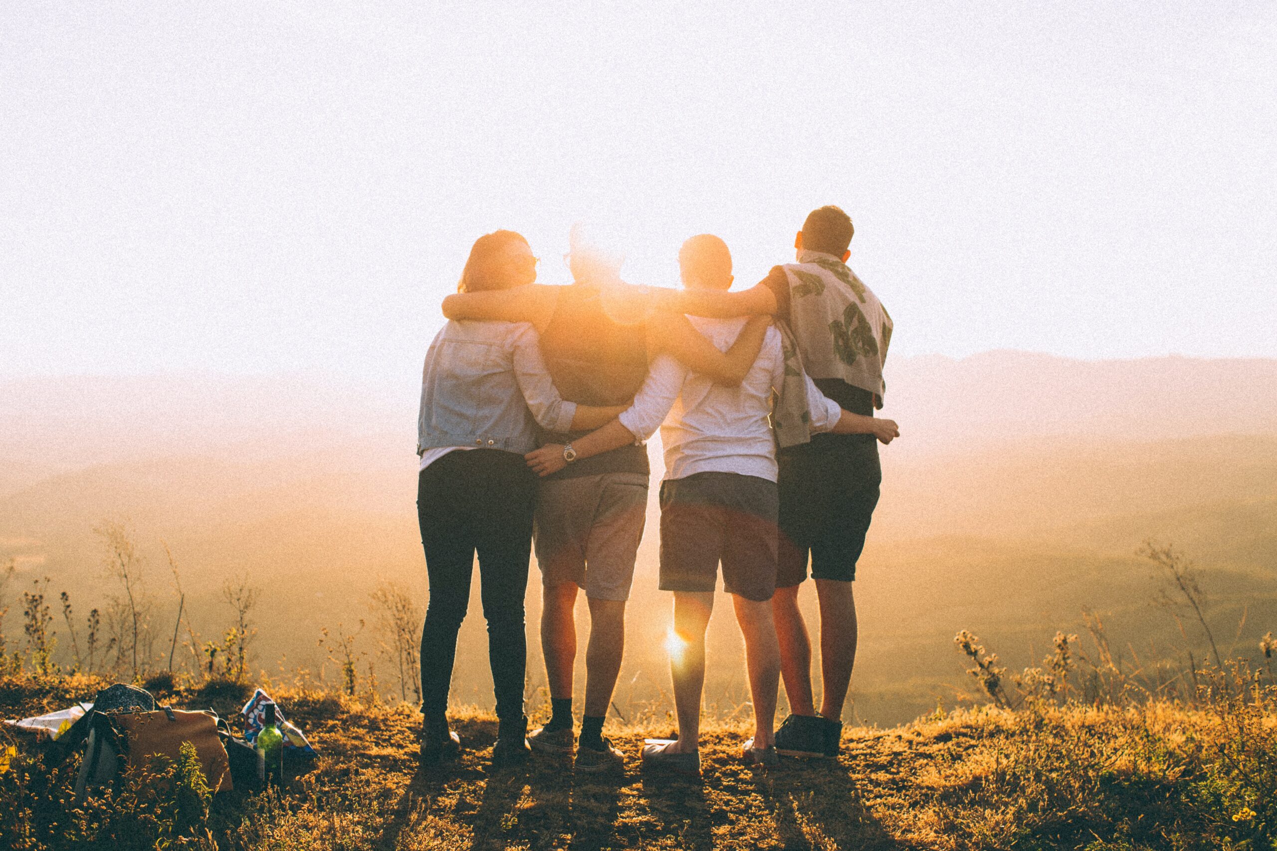 back of four people with arms around each on a mountain top watching the sun set