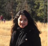Portrait of Berenice Garcia Tellez, a woman with long, dark hair wearing a black jacket, standing outside in front of a brown grassy field with scattered evergreen trees