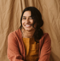 Portrait of Kiran Herbert, a woman with wavy dark hair pulled into a low ponytail, wearing a yellow-orange top and red-orange jacket, sitting in front of a warm tan fabric background
