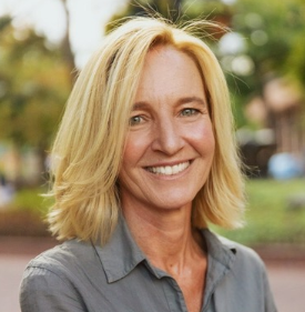 Portrait of Robyn Griggs Lawrence, a woman with shoulder-length blonde hair wearing a grey button down shirt, standing outside in front of a blurred green and brown landscape