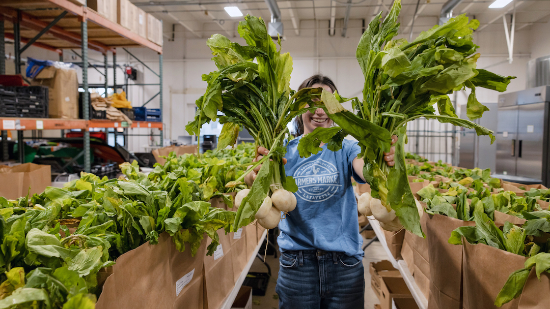 Employee in a blue t-shirt holds up green vegetables to the camera with rows of produce bags lined up in the background