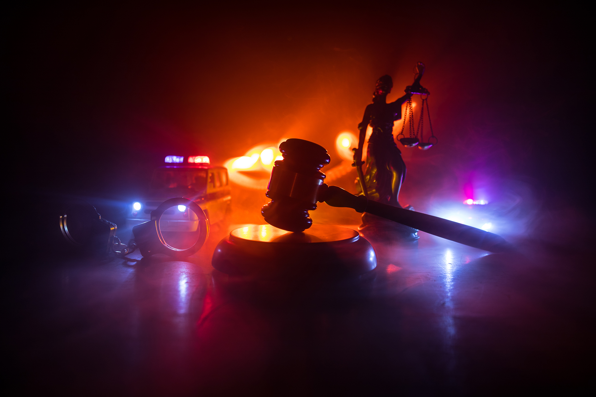 A gavel in front of a smokey background of a police car lights and a statue of a blindfolded lady holding a sword and scales.