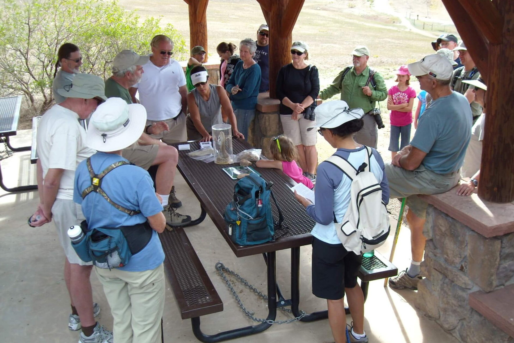 A large group of people gather around a park shelter listening to a tour guide