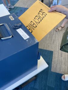 someone's hand holding an envelope marked Secrecy Envelope, which holds a ballot, and inserting envelope into ballot box
