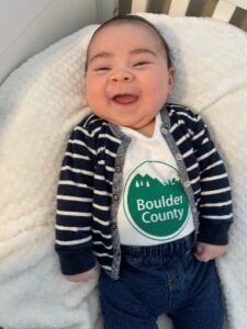 Baby laying down in crib with a big smile and a Boulder County shirt