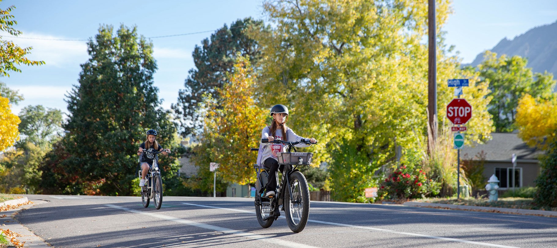 Two youth wearing helmets ride eBikes in a bike lane in Boulder County. Trees and mountains can be seen in the background.