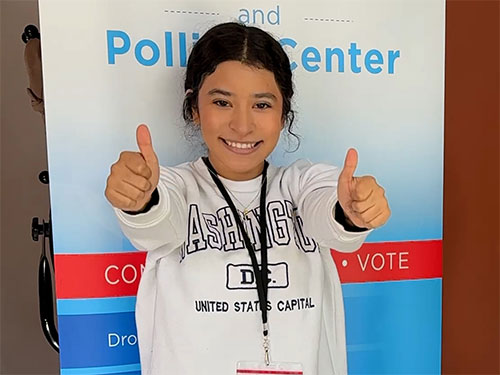 A student election judge stands in front of an elections banner and gives two thumbs up