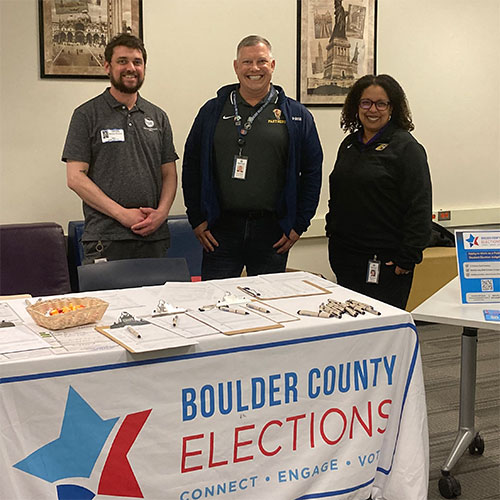 Three people at a high school voter registration event stand behind a Boulder County Elections table.