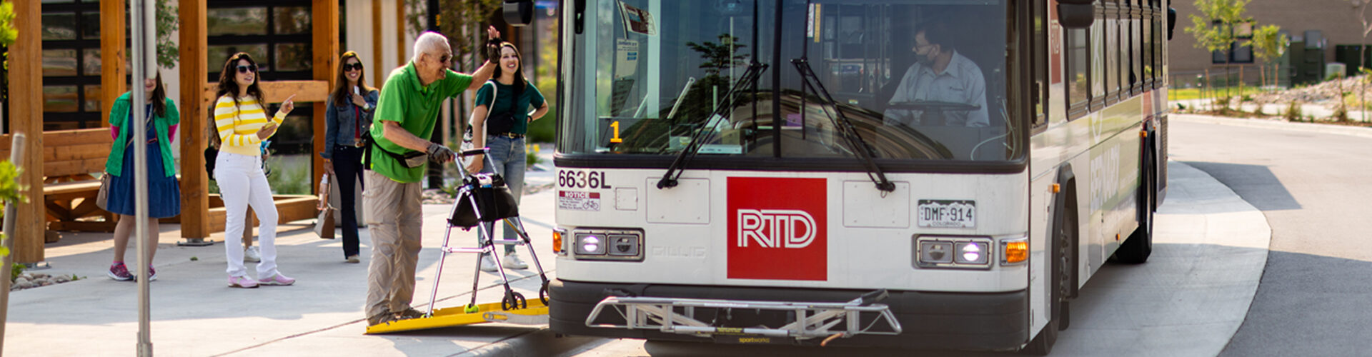 Older adult boarding an RTD bus using a walker while waving at the bus driver, with smiling onlookers.