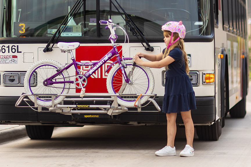 Young girl wearing pink bike helmet loading a bike on the front of an RTD bus.