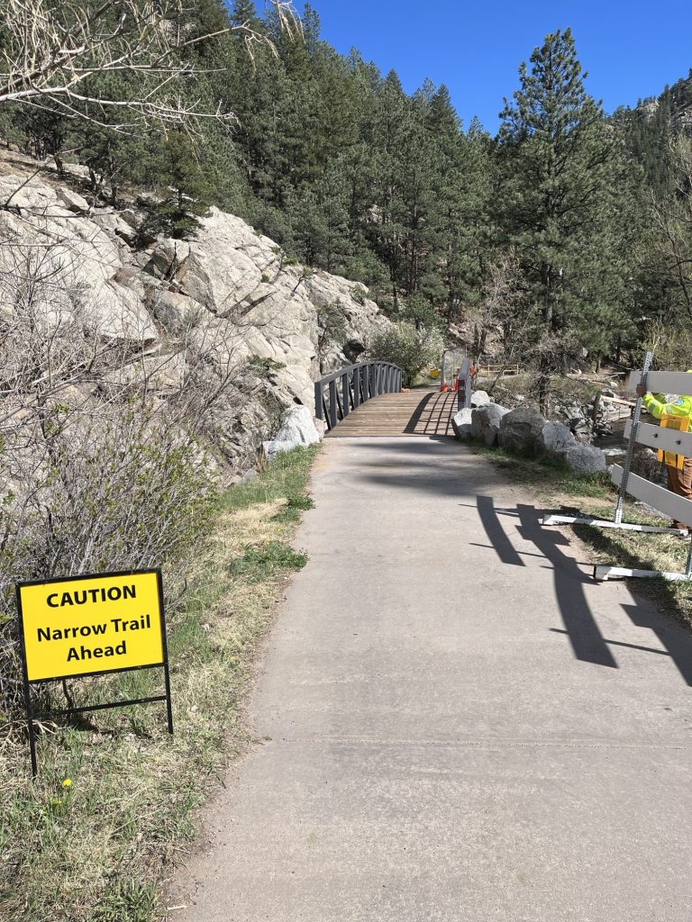 A sign next to a trail that reads Caution Narrow Trail Ahead