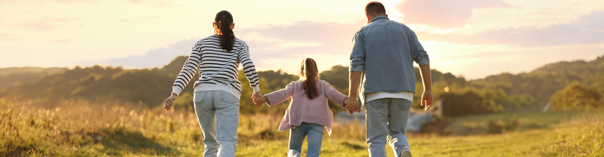 A family of three, two adults and a child, walk hand in hand through a sunlit grassy field at sunset, viewed from behind.
