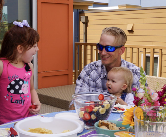 father with toddler and young child sitting on the deck at a table with food