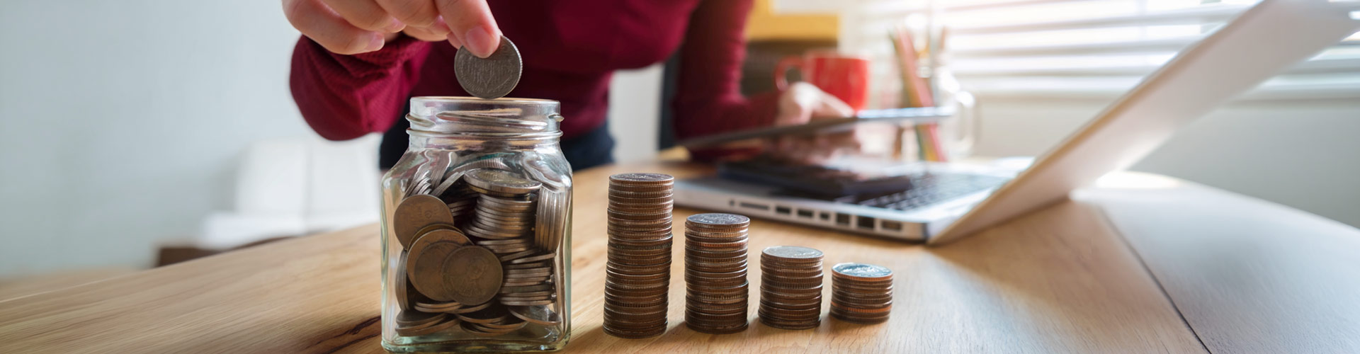 Woman placing coins into a glass jar while holding a calculator, with stacked coins arranged like a rising graph and a laptop on the desk, representing budgeting and savings.