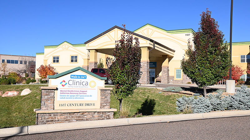 Outside view of a Clinica Family Health & Wellness building with the clinic in the background and a cement sign in front displaying ‘Walk-In 24/7’ along with the clinic name, address, and additional details