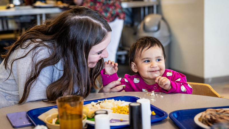 Smiling mother feeding a happy child seated at a table with a plate full of food.