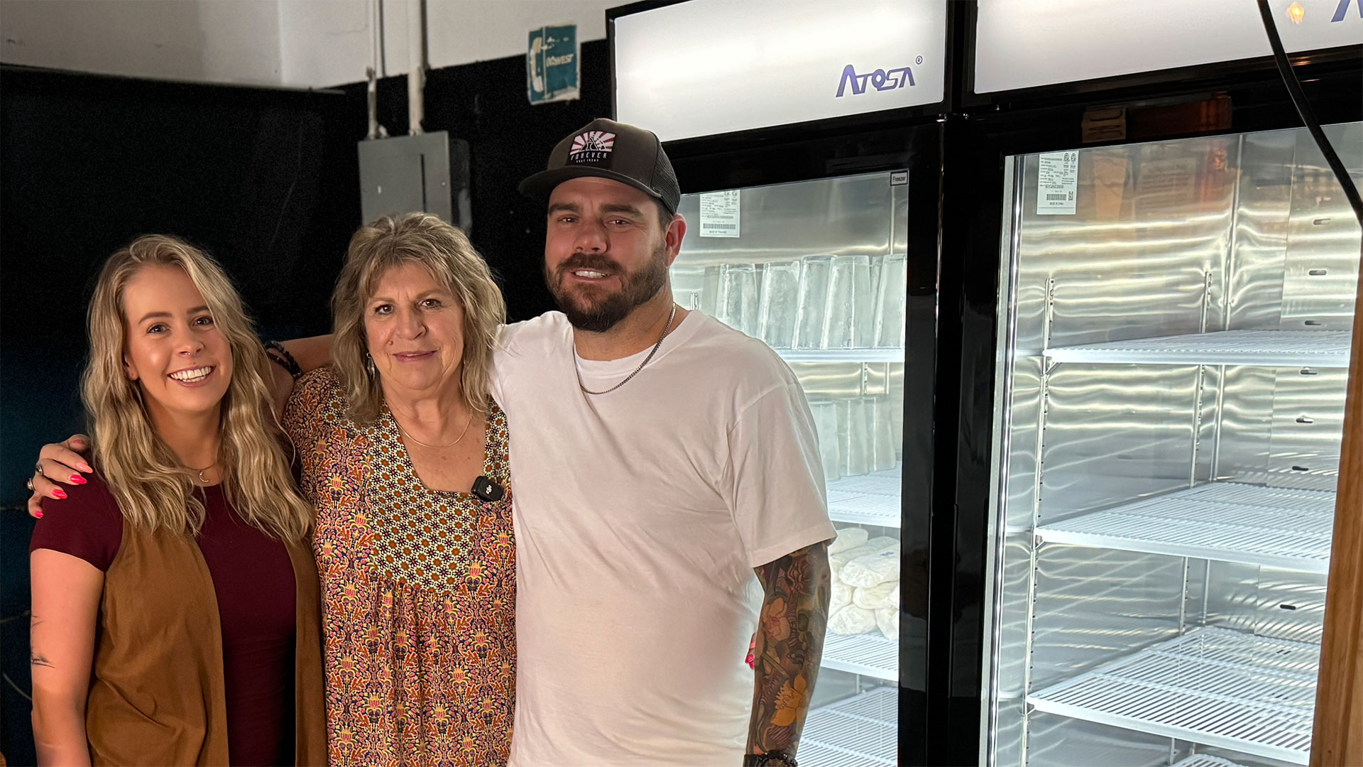 Two women and a man stand in front of a new commercial refrigerator. They are smiling and they have their arms around each other.