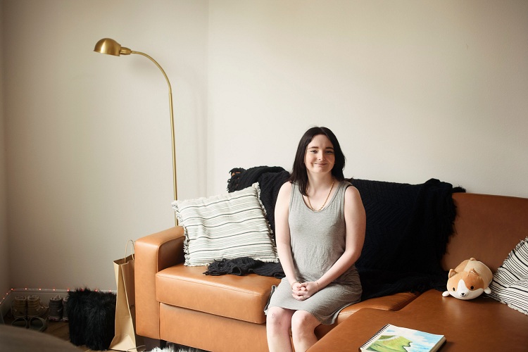 Young woman sits on a couch with her hands in her lap and a smile and her face.
