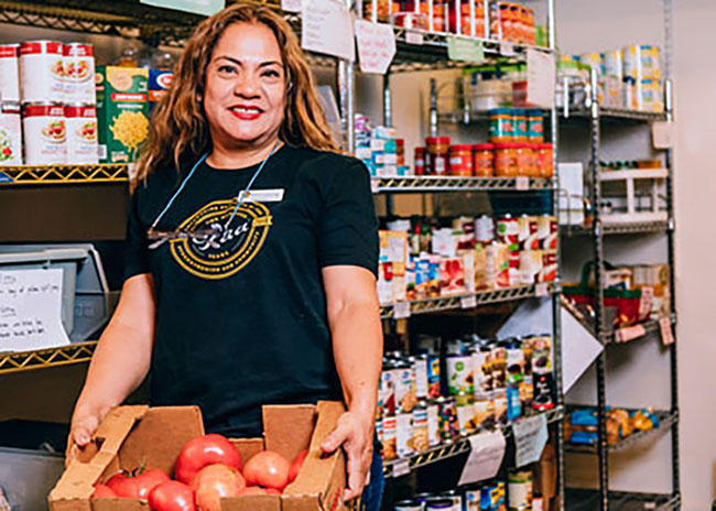 efaa-profile-photo A woman holding a box of tomatoes in a food pantry, smiling at the camera.