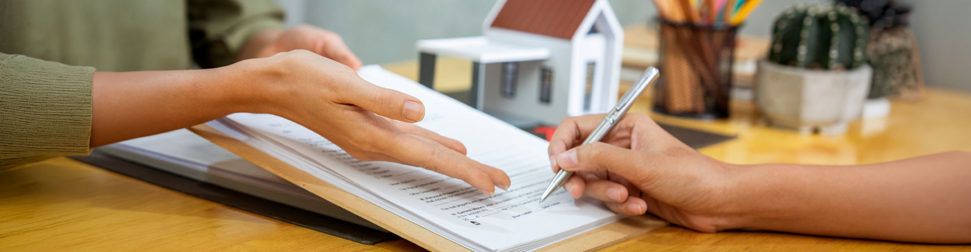 Two people review a document together. One person points to the paper while the other holds a pen to sign. A small model house, a colorful pencil holder, and a tiny cactus are on the table in the background.