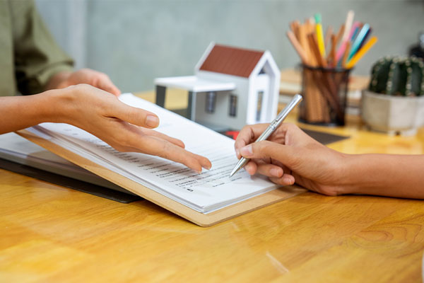 Two people review a document together. One person points to the paper while the other holds a pen to sign. A small model house, a colorful pencil holder, and a tiny cactus are on the table in the background.