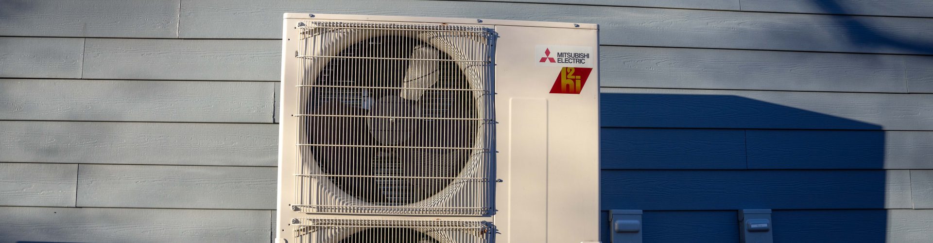 An outdoor heat pump unit sits outside a grey house with long, early-moring shadows.