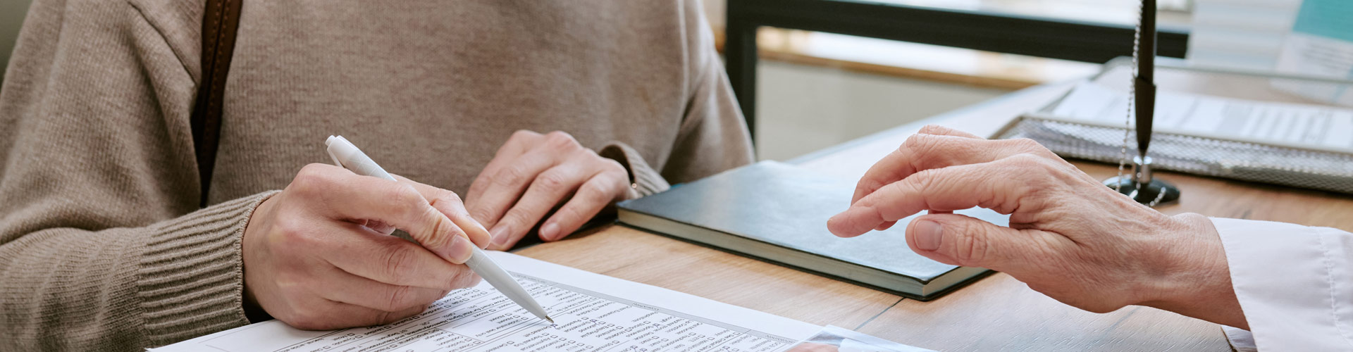 Two people sit at a desk reviewing paperwork; one writes on a document while the other points to the page.
