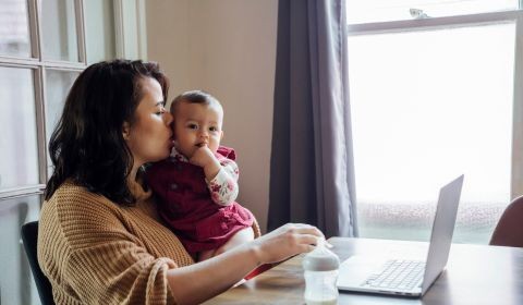 A woman sits and kisses her baby in front of a computer.